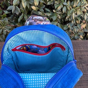 May include: Blue backpack with a zippered pocket and a blue and white checkered lining. The backpack has a red zipper and a cartoon character print on the inside.