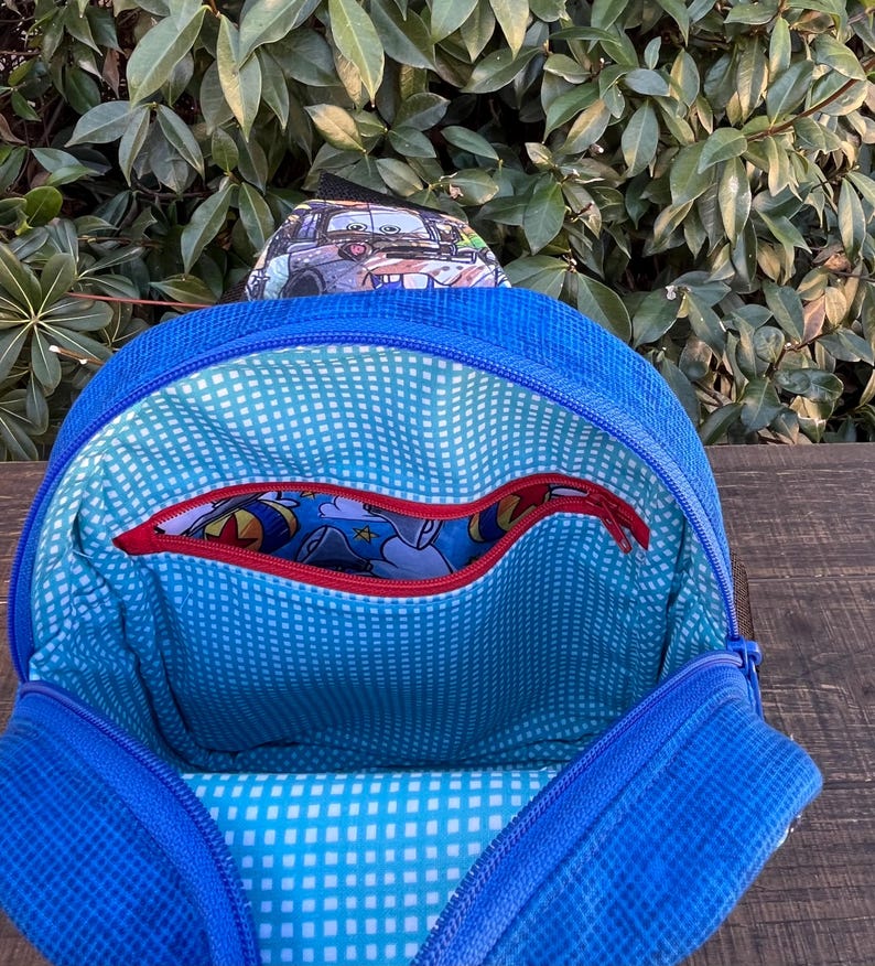 May include: Blue backpack with a zippered pocket and a blue and white checkered lining. The backpack has a red zipper and a cartoon character print on the inside.