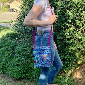 Puede incluir: Un bolso bandolera azul con un estampado colorido de personajes de dibujos animados. El bolso tiene una correa morada y m&uacute;ltiples compartimentos con cremallera. El bolso se lleva al hombro.