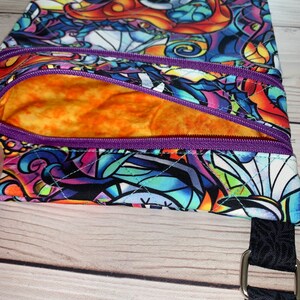 May include: A quilted crossbody bag featuring a vibrant, multi-colored stained glass pattern with character faces. The bag includes a purple zippered front pocket, an orange interior lining, and a black strap attached with a silver metal ring.