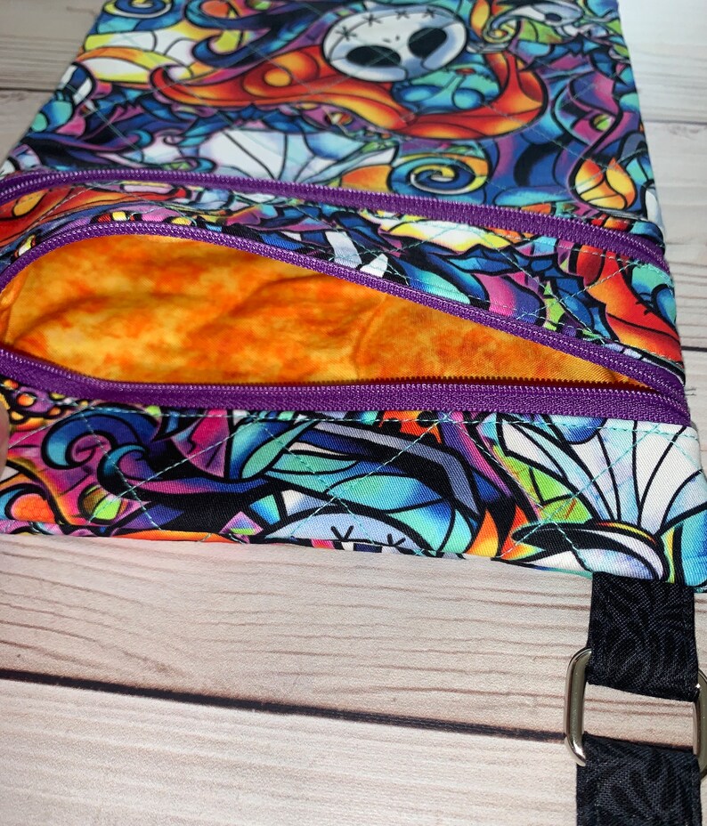 May include: A quilted crossbody bag featuring a vibrant, multi-colored stained glass pattern with character faces. The bag includes a purple zippered front pocket, an orange interior lining, and a black strap attached with a silver metal ring.