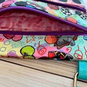 K&ouml;nnte beinhalten: Eine Umh&auml;ngetasche mit mehreren F&auml;chern und einem farbenfrohen Cartoon-Charakter-Aufdruck. Die Tasche hat einen t&uuml;rkisfarbenen Riemen und lila Rei&szlig;verschl&uuml;sse. Das Innenfutter ist ein rosa Muster. Die Tasche ist aus gestepptem Stoff gefertigt.