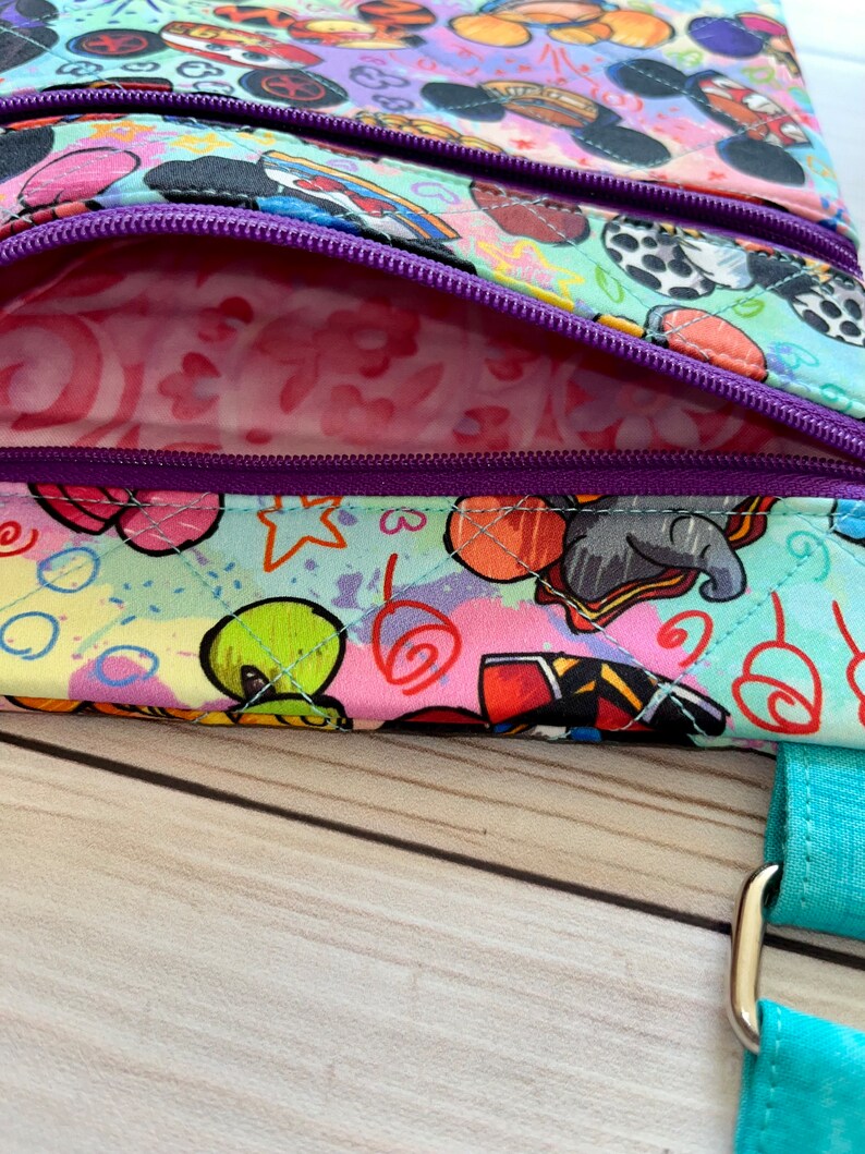 K&ouml;nnte beinhalten: Eine Umh&auml;ngetasche mit mehreren F&auml;chern und einem farbenfrohen Cartoon-Charakter-Aufdruck. Die Tasche hat einen t&uuml;rkisfarbenen Riemen und lila Rei&szlig;verschl&uuml;sse. Das Innenfutter ist ein rosa Muster. Die Tasche ist aus gestepptem Stoff gefertigt.