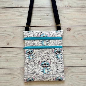 May include: A crossbody bag featuring a white fabric with a repeating blue and black sketch-style print of the character Stitch, accented by two bright turquoise horizontal zippers and a black adjustable strap with subtle white polka dots.