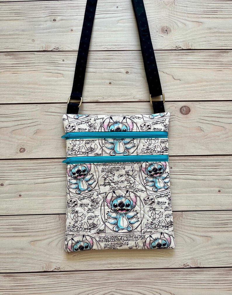 May include: A crossbody bag featuring a white fabric with a repeating blue and black sketch-style print of the character Stitch, accented by two bright turquoise horizontal zippers and a black adjustable strap with subtle white polka dots.