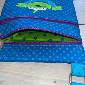 May include: Blue quilted fabric bag with a green alien character applique. The bag features a purple zipper and a blue polka dot pattern. The interior is green with blue heart shapes. A blue strap with a metal buckle is attached.