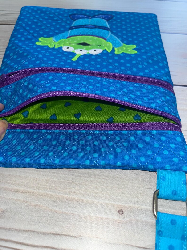 May include: Blue quilted fabric bag with a green alien character applique. The bag features a purple zipper and a blue polka dot pattern. The interior is green with blue heart shapes. A blue strap with a metal buckle is attached.
