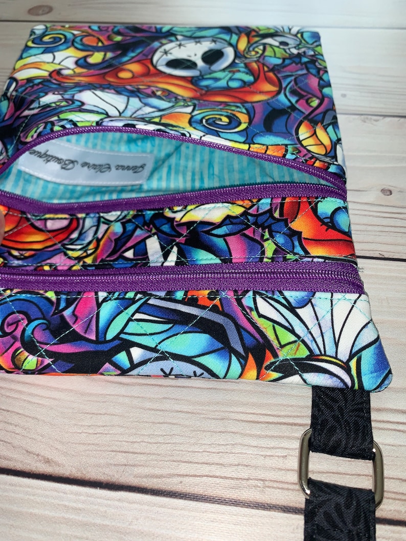 May include: A quilted fabric pouch featuring a colorful, stained-glass style print of Jack Skellington. The pouch has two purple zippered compartments and a light blue striped interior lining with a white label reading Jenna Claire Boutique.