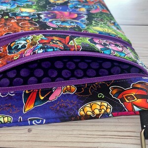 Puede incluir: Bolso bandolera multicolor con m&uacute;ltiples compartimentos con cremallera. El bolso presenta un estampado vibrante de personajes de dibujos animados con disfraces de Halloween. El interior es morado oscuro con lunares y las cremalleras son moradas.