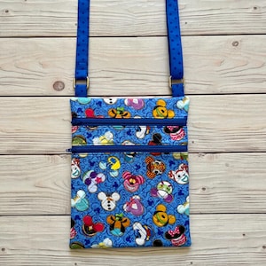 Puede incluir: Bolso bandolera azul con un estampado repetido de caras de personajes de dibujos animados. El bolso tiene dos compartimentos con cremallera y una correa azul ajustable. La correa est&aacute; sujeta con herrajes met&aacute;licos dorados.