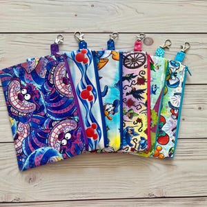May include: A collection of six rectangular fabric zipper pouches, each featuring a unique colorful cartoon-themed print and a metal swivel clasp attached to the top zipper. The designs include patterns with a purple cat, red mouse silhouettes, a duck character, a floral scene with a carriage wheel, and various other animated motifs.