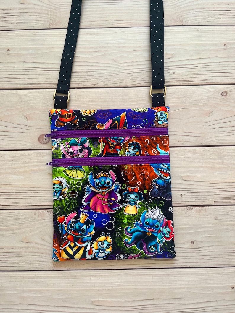 Puede incluir: Bolso bandolera rectangular con estampado de personajes de dibujos animados coloridos. El bolso tiene una correa negra con lunares blancos, tres compartimentos con cremallera morada y herrajes dorados. El estampado incluye varios personajes con disfraces.