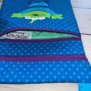 May include: A blue crossbody bag with a quilted pattern and a cartoon alien design. The bag features a purple zipper and a blue strap. The alien is green with three eyes and is wearing a blue outfit. The bag has the text "Jennaclair Boutique".