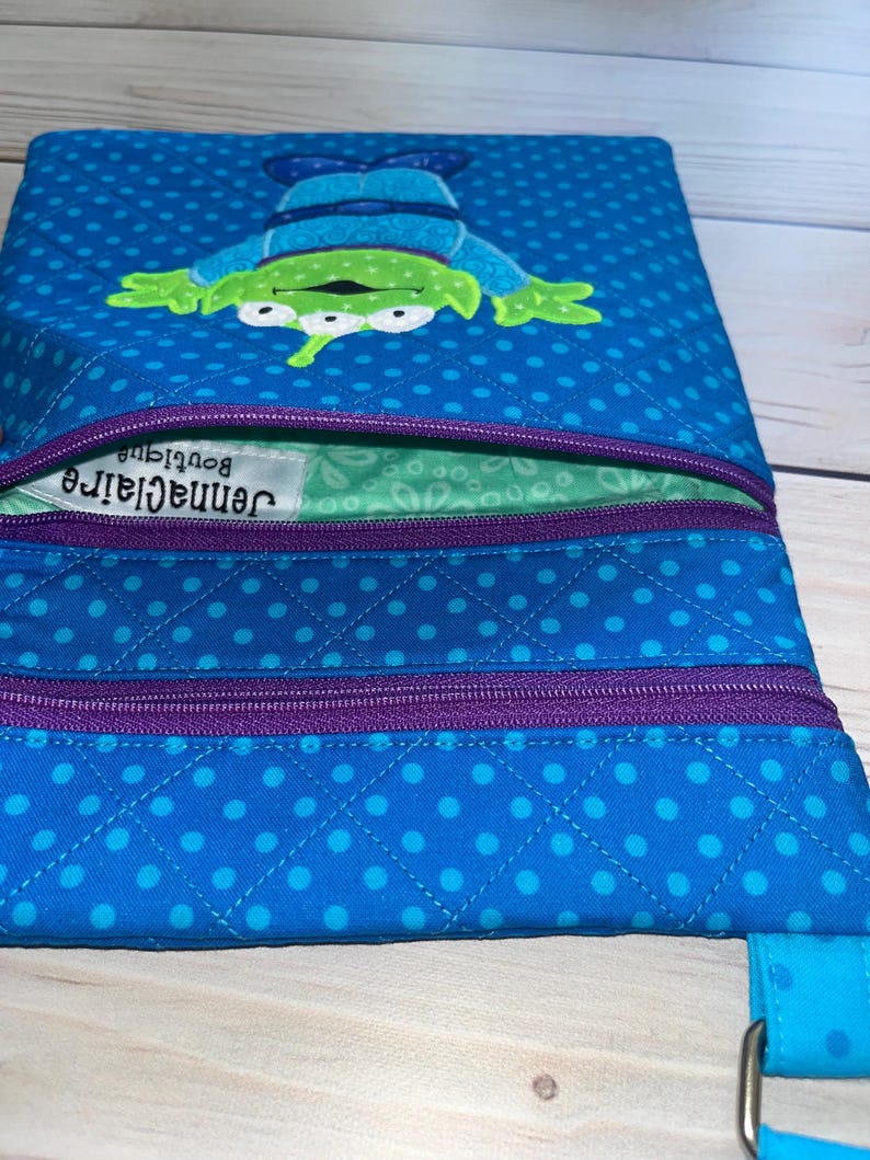 May include: A blue crossbody bag with a quilted pattern and a cartoon alien design. The bag features a purple zipper and a blue strap. The alien is green with three eyes and is wearing a blue outfit. The bag has the text "Jennaclair Boutique".