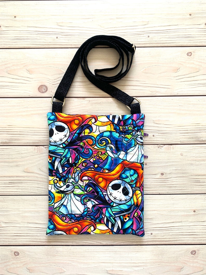 May include: A quilted crossbody bag featuring a vibrant, stained-glass style print of characters from The Nightmare Before Christmas, including Jack Skellington and Sally. The bag has a black adjustable strap and a rectangular, stitched fabric body.