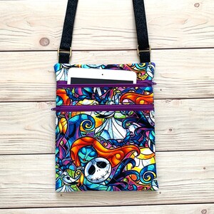 May include: A quilted crossbody bag featuring a vibrant, multicolored print of characters from The Nightmare Before Christmas. The bag has two purple zippered compartments and a black adjustable strap with a small tablet tucked inside the top pocket.