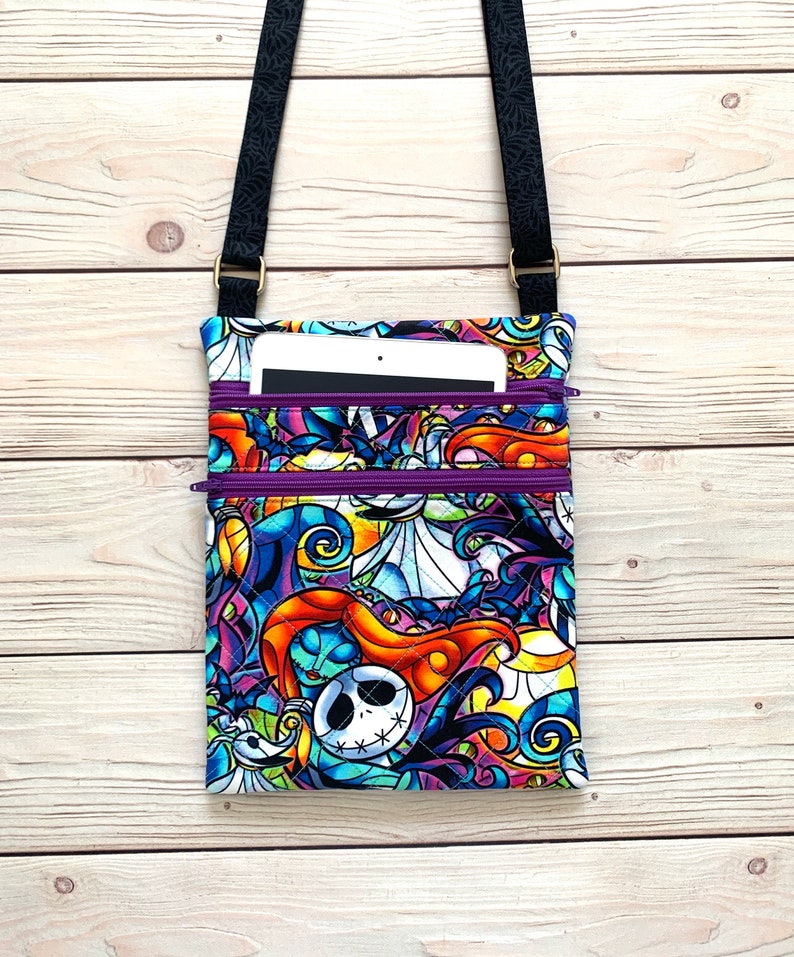 May include: A quilted crossbody bag featuring a vibrant, multicolored print of characters from The Nightmare Before Christmas. The bag has two purple zippered compartments and a black adjustable strap with a small tablet tucked inside the top pocket.