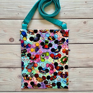 K&ouml;nnte beinhalten: Gesteppte Umh&auml;ngetasche mit einem farbenfrohen Muster aus Mickey Mouse-K&ouml;pfen und anderen Zeichentrickfiguren. Die Tasche hat einen verstellbaren, t&uuml;rkisfarbenen Riemen und ist aus Stoff. Der Hintergrund ist eine helle Holzoberfl&auml;che.