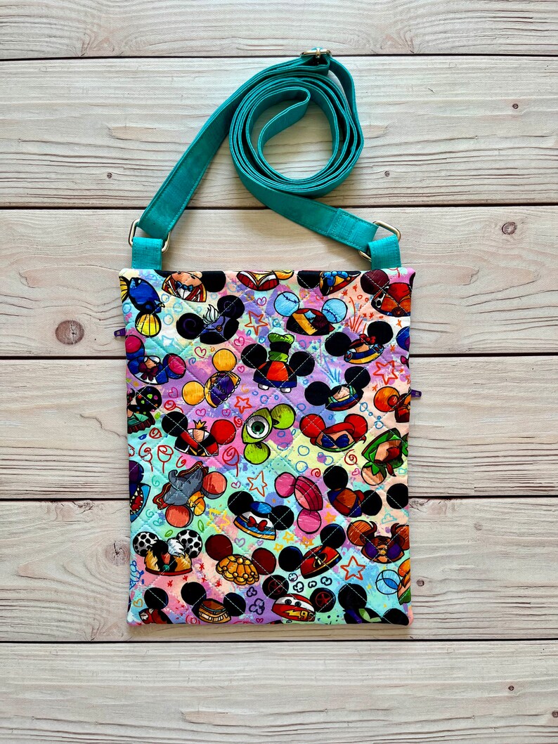 K&ouml;nnte beinhalten: Gesteppte Umh&auml;ngetasche mit einem farbenfrohen Muster aus Mickey Mouse-K&ouml;pfen und anderen Zeichentrickfiguren. Die Tasche hat einen verstellbaren, t&uuml;rkisfarbenen Riemen und ist aus Stoff. Der Hintergrund ist eine helle Holzoberfl&auml;che.