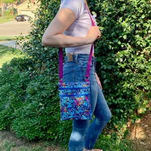 May include: A rectangular crossbody bag featuring a vibrant blue and purple cartoon character print with two horizontal zippered compartments and bright blue zippers. The bag has a purple strap with a small white polka dot pattern attached by metal rings.