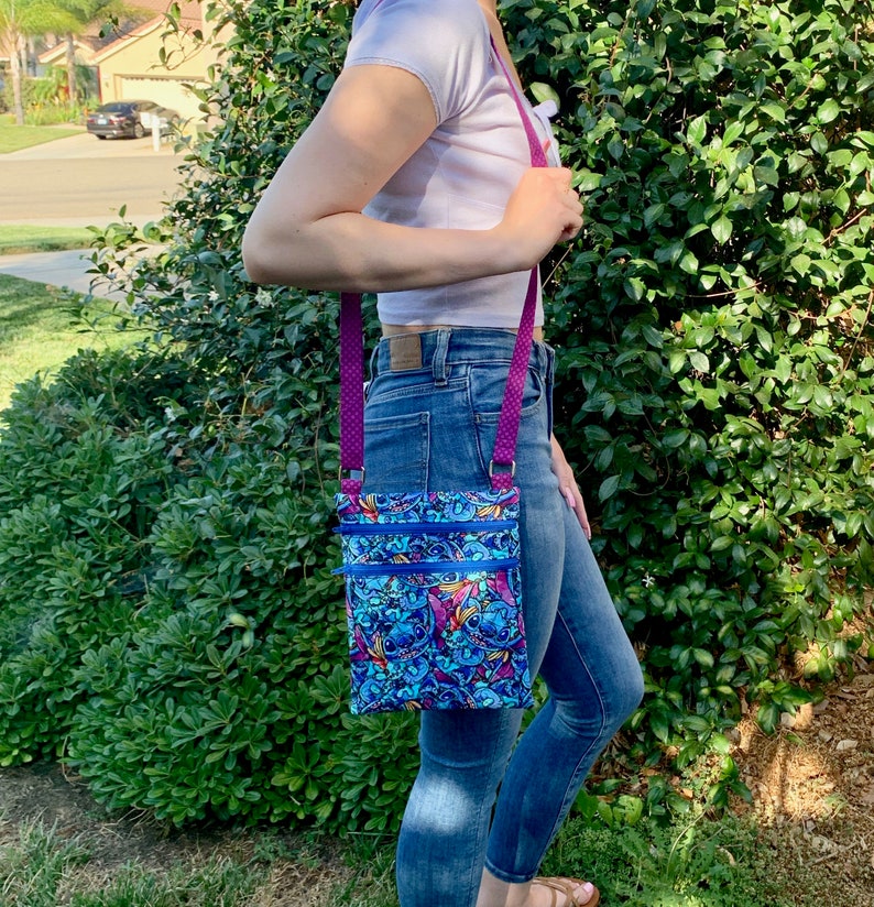May include: A rectangular crossbody bag featuring a vibrant blue and purple cartoon character print with two horizontal zippered compartments and bright blue zippers. The bag has a purple strap with a small white polka dot pattern attached by metal rings.
