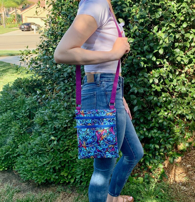 May include: A rectangular crossbody bag featuring a vibrant blue and purple pattern of the character Stitch. The bag has two horizontal zippered compartments with bright blue zips and a long, adjustable purple strap with a small white dot pattern.