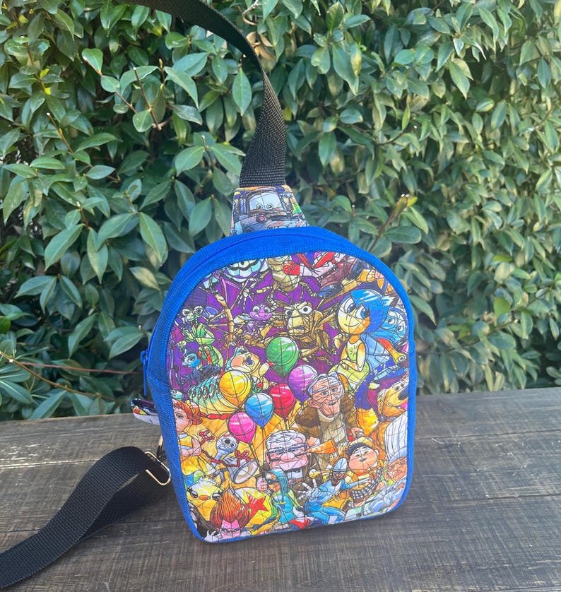 May include: A blue and black crossbody bag with a colorful print of various cartoon characters from popular animated movies. The print includes characters like Buzz Lightyear, Woody, Mike Wazowski, Sulley, and more.