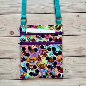 K&ouml;nnte beinhalten: Bunte Umh&auml;ngetasche mit einem Muster aus Zeichentrickfigurenk&ouml;pfen. Die Tasche hat einen t&uuml;rkisfarbenen Riemen und lila Rei&szlig;verschl&uuml;sse. Ein wei&szlig;es Tablet ist in der oberen Tasche sichtbar. Die Tasche ist aus Stoff.