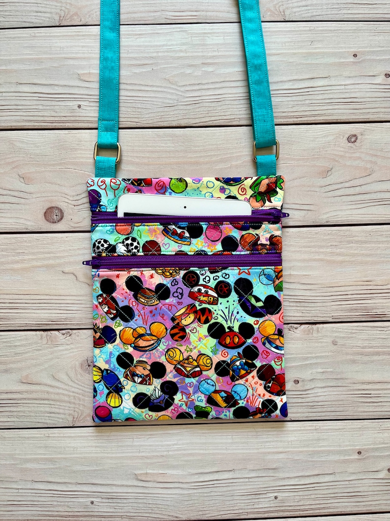 K&ouml;nnte beinhalten: Bunte Umh&auml;ngetasche mit einem Muster aus Zeichentrickfigurenk&ouml;pfen. Die Tasche hat einen t&uuml;rkisfarbenen Riemen und lila Rei&szlig;verschl&uuml;sse. Ein wei&szlig;es Tablet ist in der oberen Tasche sichtbar. Die Tasche ist aus Stoff.