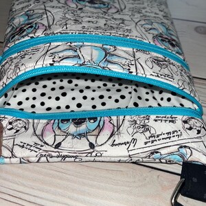 May include: A quilted crossbody bag featuring a white fabric with a blue and black sketch-style print of the character Stitch, accented by bright turquoise zips. The interior lining is white with black polka dots and a black strap is attached.