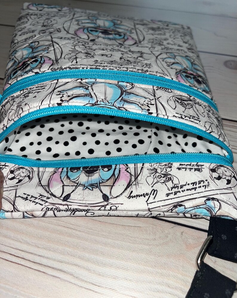 May include: A quilted crossbody bag featuring a white fabric with a blue and black sketch-style print of the character Stitch, accented by bright turquoise zips. The interior lining is white with black polka dots and a black strap is attached.