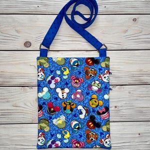 Puede incluir: Un bolso bandolera azul con un estampado repetido de caras de personajes de dibujos animados. El bolso tiene una correa azul con lunares blancos y un cierre de cremallera. El bolso es de tela.