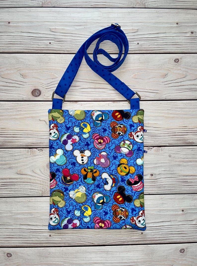 Puede incluir: Un bolso bandolera azul con un estampado repetido de caras de personajes de dibujos animados. El bolso tiene una correa azul con lunares blancos y un cierre de cremallera. El bolso es de tela.