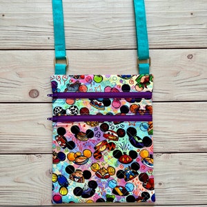 K&ouml;nnte beinhalten: Eine farbenfrohe Umh&auml;ngetasche mit einem lebendigen Muster aus Zeichentrickfigurenk&ouml;pfen und anderen Grafiken. Die Tasche hat zwei Rei&szlig;verschlussf&auml;cher mit lila Rei&szlig;verschl&uuml;ssen und einen verstellbaren, t&uuml;rkisfarbenen Riemen. Die Tasche ist aus gestepptem Stoff gefertigt.