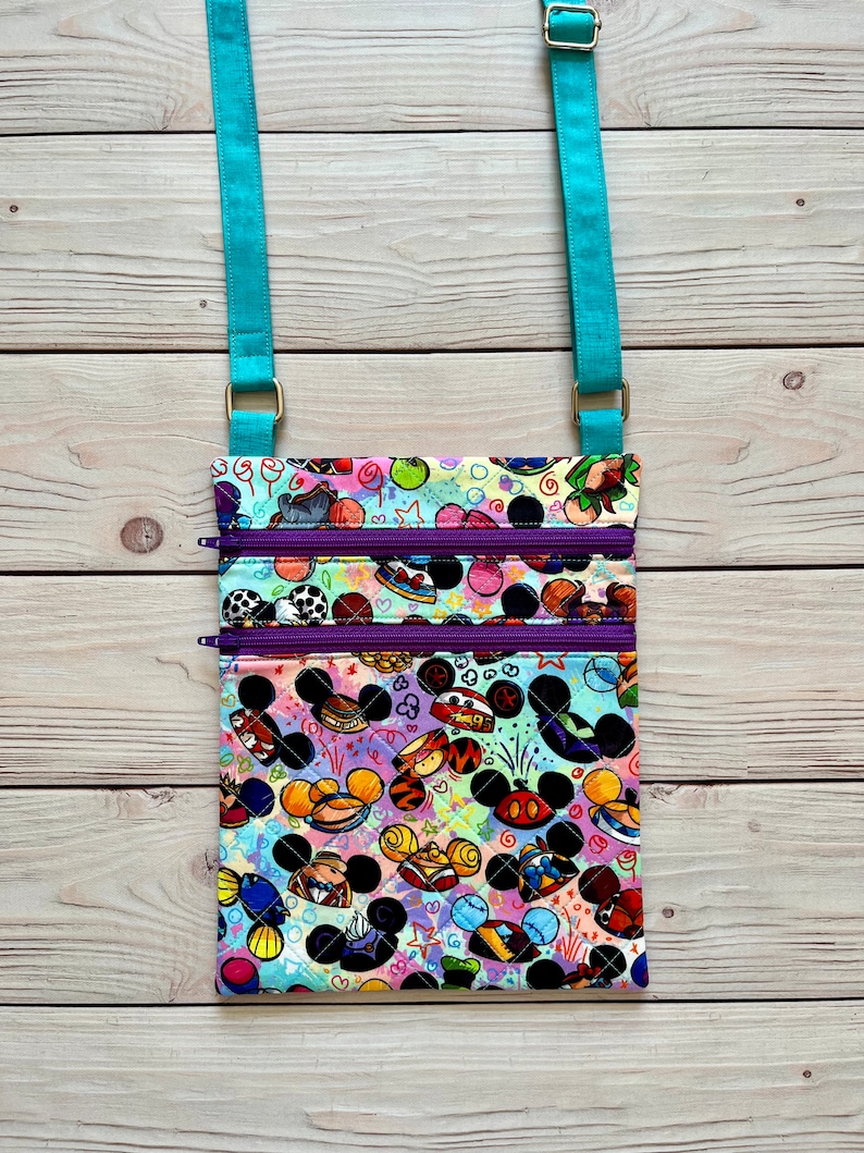 K&ouml;nnte beinhalten: Eine farbenfrohe Umh&auml;ngetasche mit einem lebendigen Muster aus Zeichentrickfigurenk&ouml;pfen und anderen Grafiken. Die Tasche hat zwei Rei&szlig;verschlussf&auml;cher mit lila Rei&szlig;verschl&uuml;ssen und einen verstellbaren, t&uuml;rkisfarbenen Riemen. Die Tasche ist aus gestepptem Stoff gefertigt.