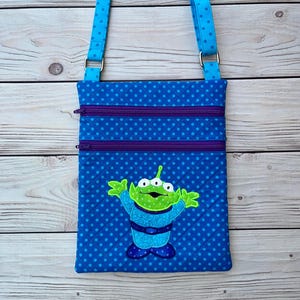 May include: Blue crossbody bag with a repeating polka dot pattern and a green alien applique. The bag has two purple zippered pockets and a blue adjustable strap. The alien has three eyes and is wearing a blue outfit.