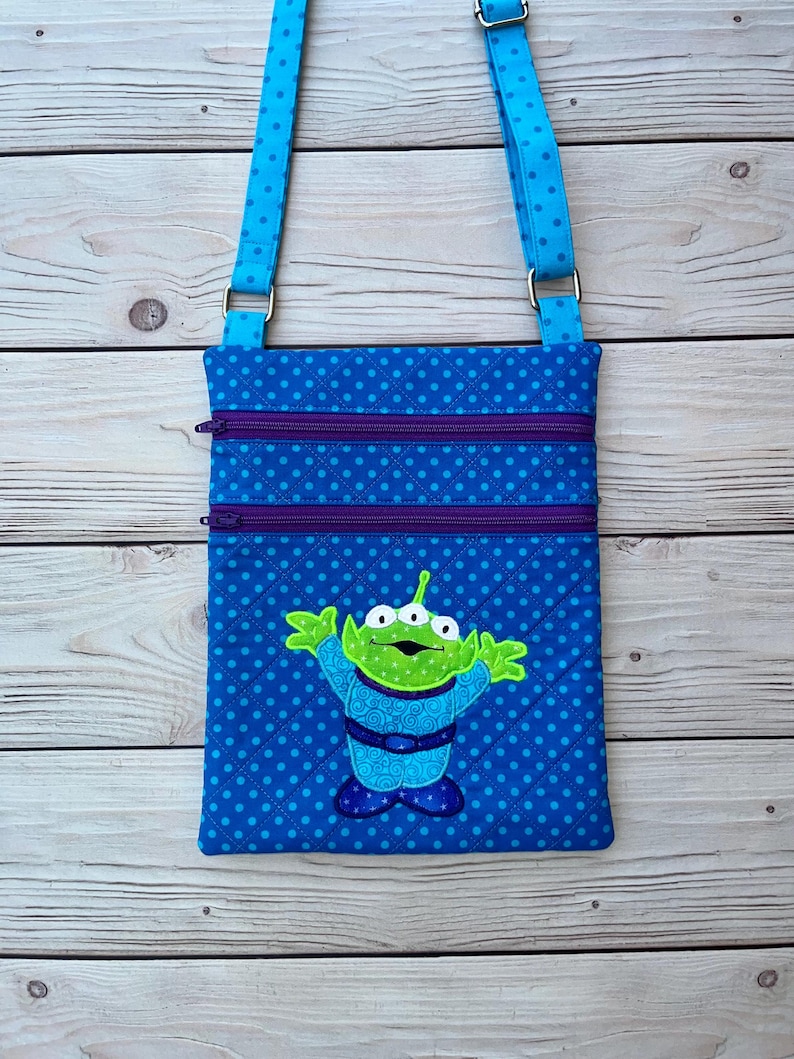 May include: Blue crossbody bag with a repeating polka dot pattern and a green alien applique. The bag has two purple zippered pockets and a blue adjustable strap. The alien has three eyes and is wearing a blue outfit.