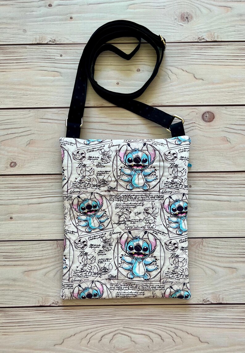 May include: A rectangular crossbody bag featuring a light-coloured fabric with a repeating print of the blue character Stitch from Lilo & Stitch, surrounded by scientific sketches, diagrams, and text including the word Warning.