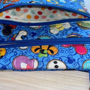 Puede incluir: Bolso de tela azul con un estampado repetido de caras de personajes de dibujos animados. El bolso tiene dos compartimentos con cremallera y cremalleras azules. El forro interior es blanco con lunares de colores.