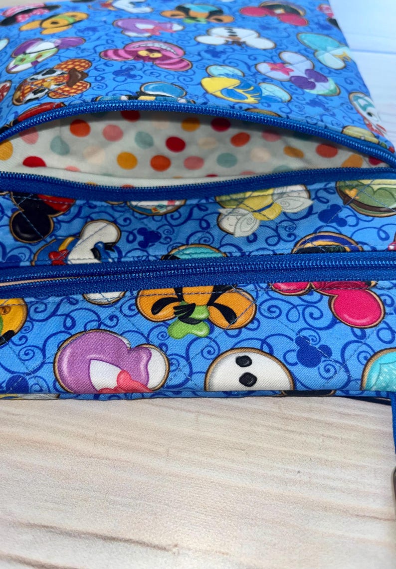 Puede incluir: Bolso de tela azul con un estampado repetido de caras de personajes de dibujos animados. El bolso tiene dos compartimentos con cremallera y cremalleras azules. El forro interior es blanco con lunares de colores.