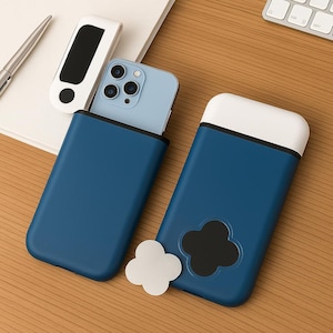 Op de afbeelding: Twee blauwe telefoonhoesjes met witte accenten en een klavervormig ontwerp. Een lichtblauwe telefoon is gedeeltelijk in een hoesje geplaatst. Een witte klavervormige sticker ligt naast de hoesjes. Een wit en zwart accessoire.