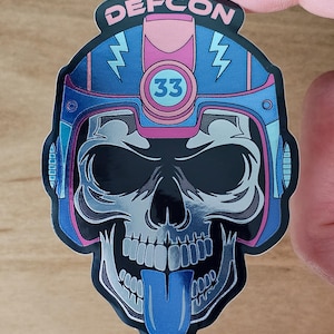 May include: A sticker featuring a skull wearing a blue and pink helmet with the text "DEFCON" at the top. The skull has a blue tongue sticking out and the number "33" is in a circle.