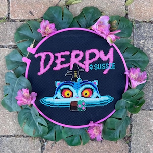 May include: A cross-stitch embroidery hoop with the word "DERPY" in pink and "SUSSIE" in teal. The design features a cartoon cat. The hoop is framed by green leaves and pink flowers.