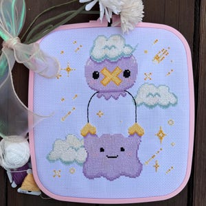 May include: Cross-stitch art featuring a purple creature holding a cloud, set against a light blue background with stars and clouds. The design is framed in a pink square, with white flowers and a ribbon on the side.