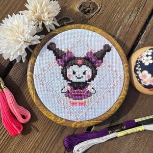 May include: A cross-stitch embroidery hoop with a white background and a purple and black cartoon character with a pink dress and white snowflakes. The hoop is made of wood with a brown stain.