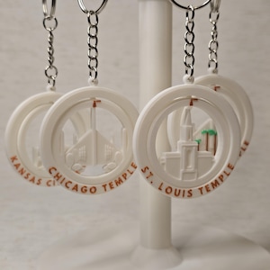 May include: Four white keychains with silver chains. Each keychain features a 3D printed design of a temple with the city and state name printed below. The keychains are: Kansas City Temple, Chicago Temple, St. Louis Temple, and a fourth temple with an unclear name.