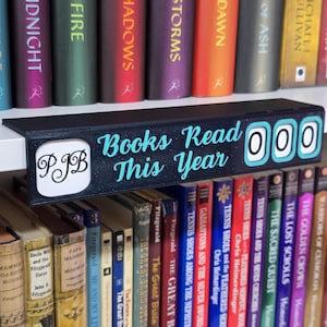 May include: A black shelf-sitter with the text "Books Read This Year" in teal. It has a counter with three white number wheels. The shelf-sitter also has the initials "PJB" in a white square. Books are visible on the shelves.