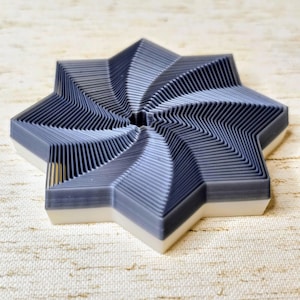 May include: A gray and white star-shaped object with a layered, geometric design. The star has eight points and a spiral pattern in the center. The base is white, and the top is gray.