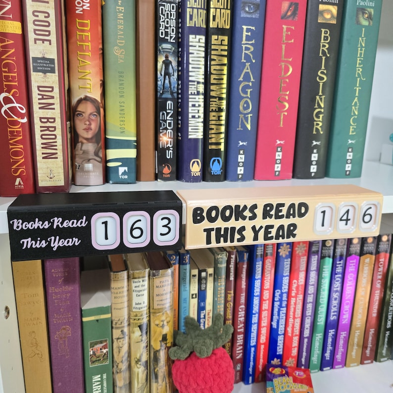May include: A bookshelf filled with colorful books, with two wooden blocks displaying the text "Books Read This Year" and the numbers 163 and 146. A crocheted strawberry sits on the shelf.
