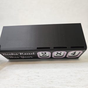 May include: A black rectangular book counter with the text "Books Read This Year". The counter displays the number "284" in white digits on a light purple background. The item is designed to track the number of books read.